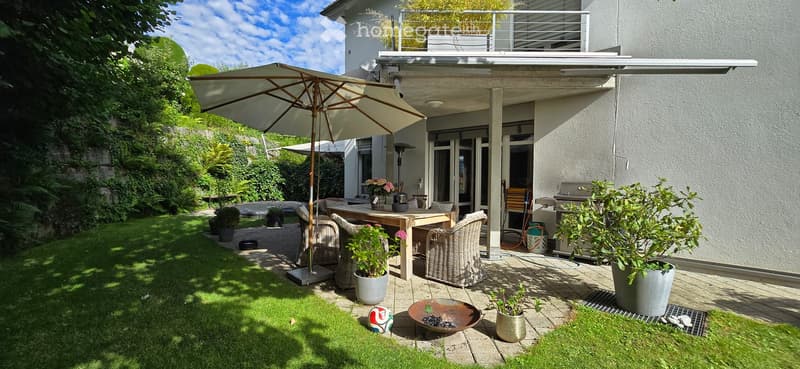 Well-maintained garden with a patio area, outdoor dining table, wicker chairs, umbrella, potted plants, and a modern house with a balcony and covered patio.