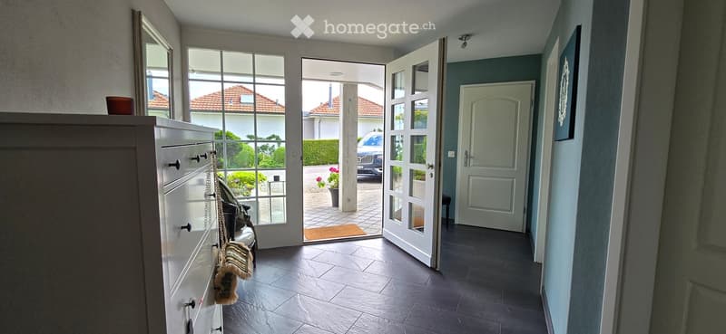 Bright hallway with dark tiled floor, white walls, large windows, and an open door leading to a patio area with outdoor furniture and a parked car visible.