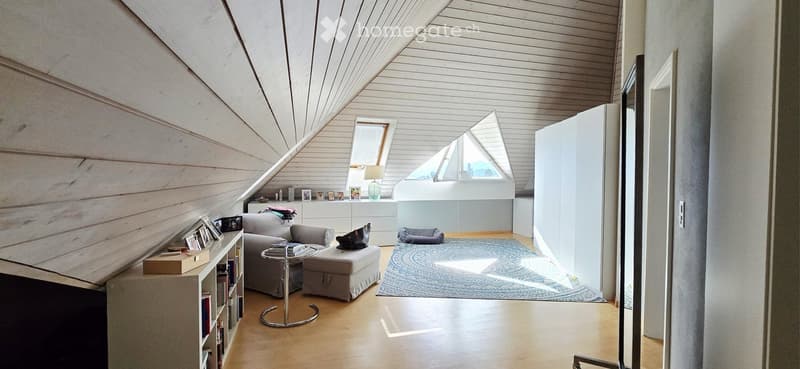 Bright, modern living room with wooden floors, white walls, and slanted wooden ceiling. Features a grey sofa, ottoman, coffee table, bookshelf, and a large window letting in natural light.