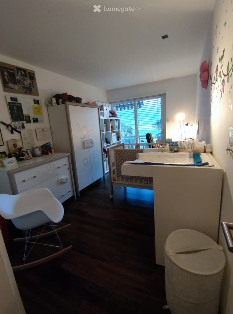 A well-lit bedroom with a wooden floor, a white rocking chair, a wooden cradle, a cabinet, a shelf, a desk with various items, and a trash bin.