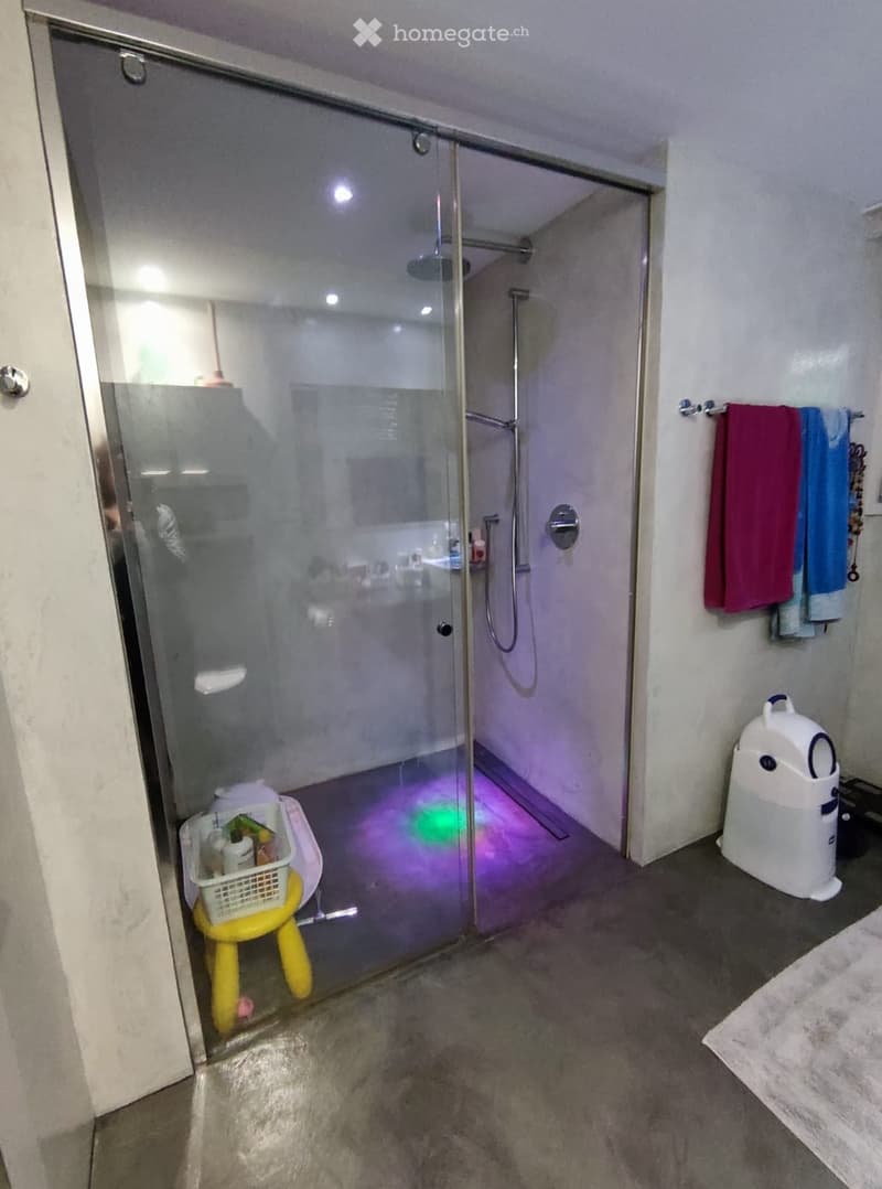 Modern bathroom with a glass-enclosed shower stall, towel rack with towels, small yellow stool with a basket, and a white trash bin.