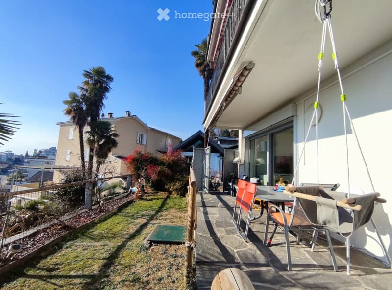 Balcony with a swing chair, table, and chairs, overlooking a garden with palm trees and a neighboring house.