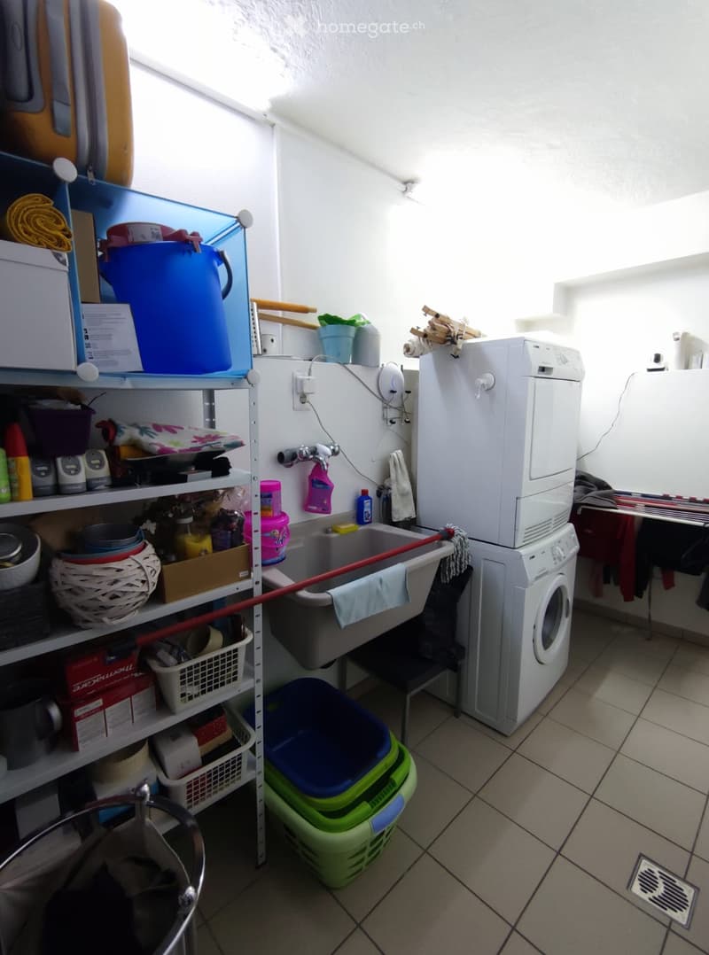 A small utility room with a washing machine, refrigerator, sink, shelves filled with cleaning supplies, and laundry items.