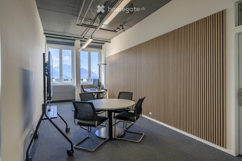 Modern office space with a round table, chairs, a large screen on a stand, a desk with a computer, and large windows offering a mountain view.