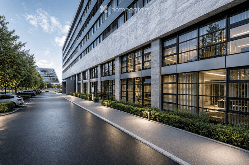 Modern multi-story building with large glass windows and a clean, contemporary design. The building is surrounded by a landscaped area with trees and shrubs, and there is a paved walkway leading to the entrance. Several parked cars are visible in the fore