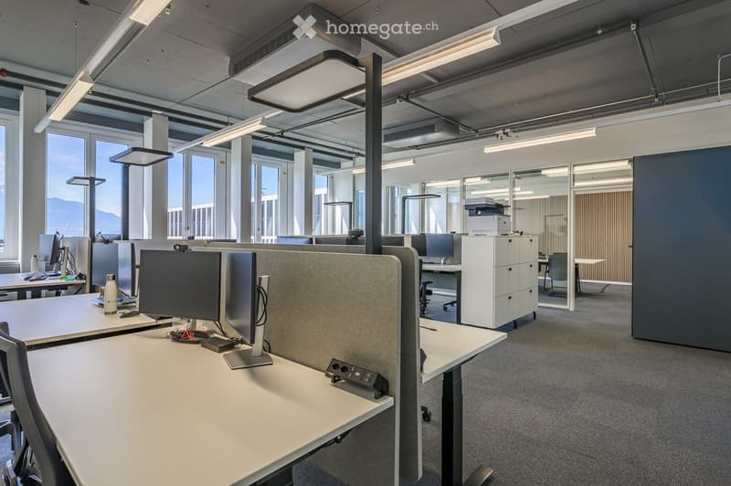 Modern office space with desks, chairs, computer monitors, and a printer. Large windows provide natural light, and the room features exposed ceiling beams and modern lighting.