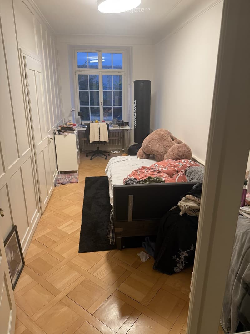 Cozy bedroom with wooden herringbone flooring, large window, bed with colorful bedding, desk with chair, storage cabinet, punching bag, and picture frame on wall.