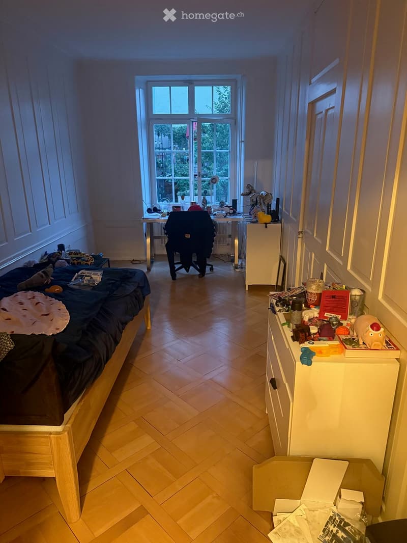 A small, well-lit bedroom with a wooden bed, desk, chair, and cabinet. The room features wooden flooring and large windows with a view of greenery.