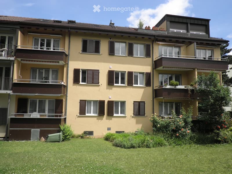 Four-story residential building with beige walls, multiple windows with shutters, balconies, and a well-maintained lawn in front.