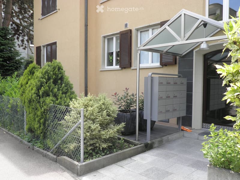 Modern building entrance with a covered porch, mailboxes, and small garden area with shrubs and potted plants.