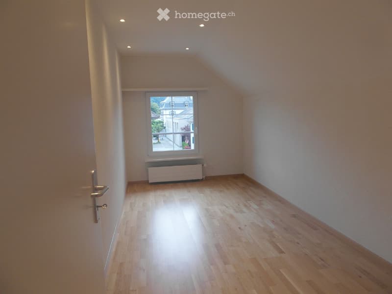 Empty room with wooden flooring, white walls, and a single window with a radiator below it. The room has a slanted ceiling typical of an attic space.