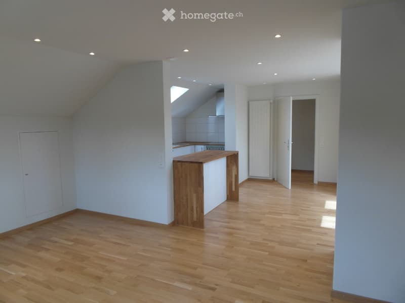 Empty room with wooden flooring, white walls, and a small kitchen area visible in the background. No furniture or equipment is present.