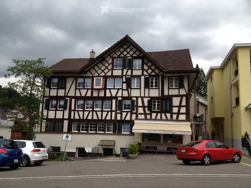 2 story building, beige paint, brown roof, windows with shutters, ground floor awning, parking area