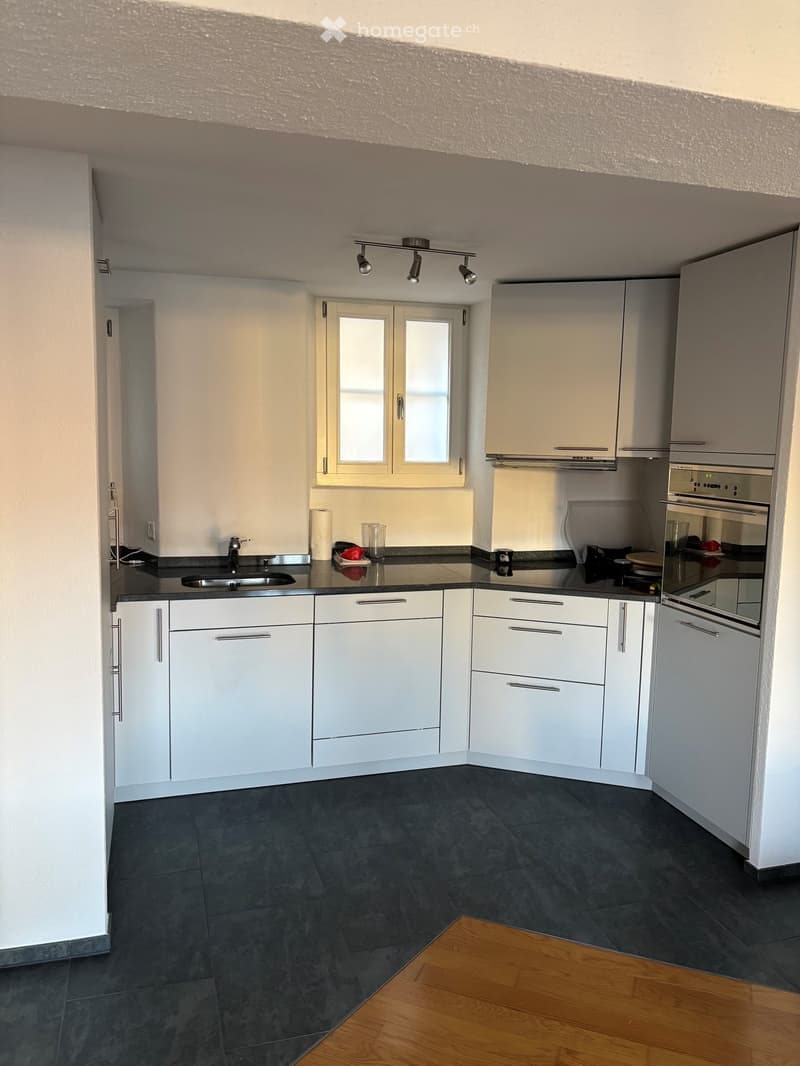Modern kitchen with white cabinets, dark countertops, and a stainless steel sink. Features include an oven, a window, and a light fixture above the sink.