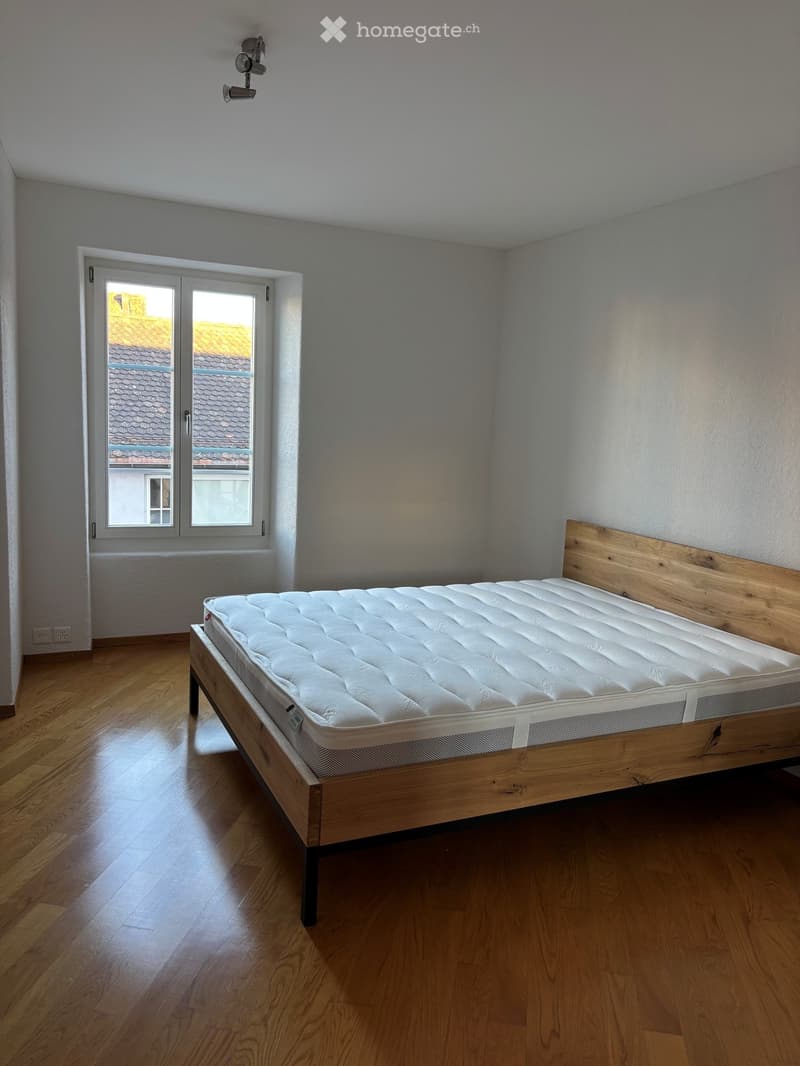 Minimalist bedroom with wooden floor, white walls, large window with view of neighboring roof, and a single bed with white mattress and wooden frame.