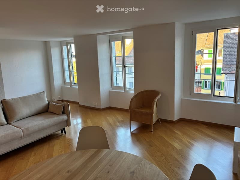 Bright living room with wooden flooring, large windows, a sofa, a wicker chair, and a wooden dining table.