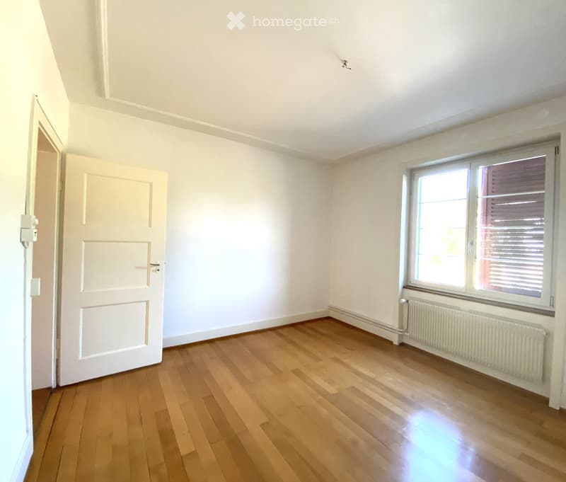 Empty room with wooden flooring, white walls, a window with blinds, and a radiator below it. The door is open, revealing a hallway.