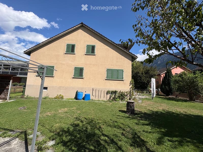Single-story house with beige siding, green shutters, and a well-maintained lawn in front. The house features a small balcony on the left side and is surrounded by trees and mountains in the background.
