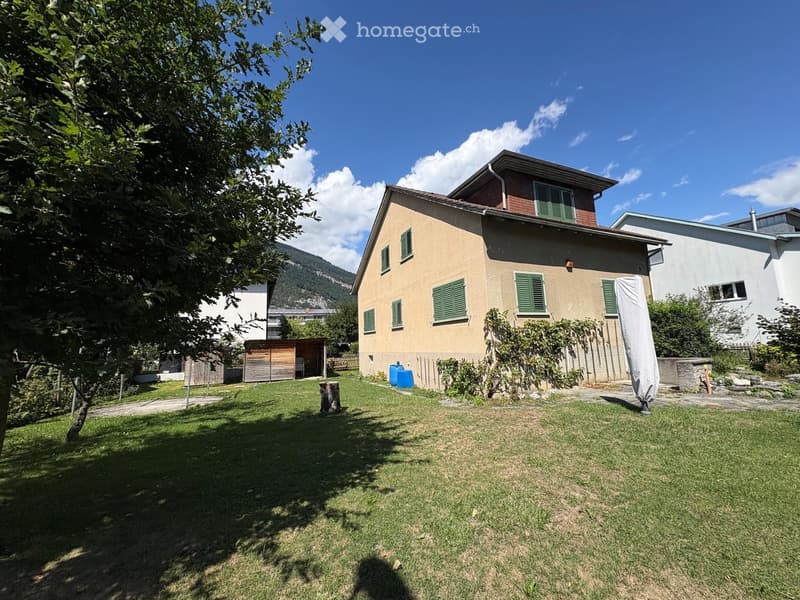 Two-story house with beige walls, green shutters, and a well-maintained lawn in front. The house features a small wooden shed and a covered patio area.