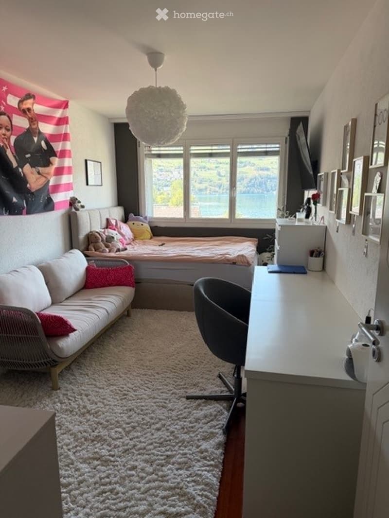 Cozy studio with a bed, sofa, desk, and large window offering a scenic view. Decor includes a fluffy white rug, colorful pillows, and framed pictures on the wall.