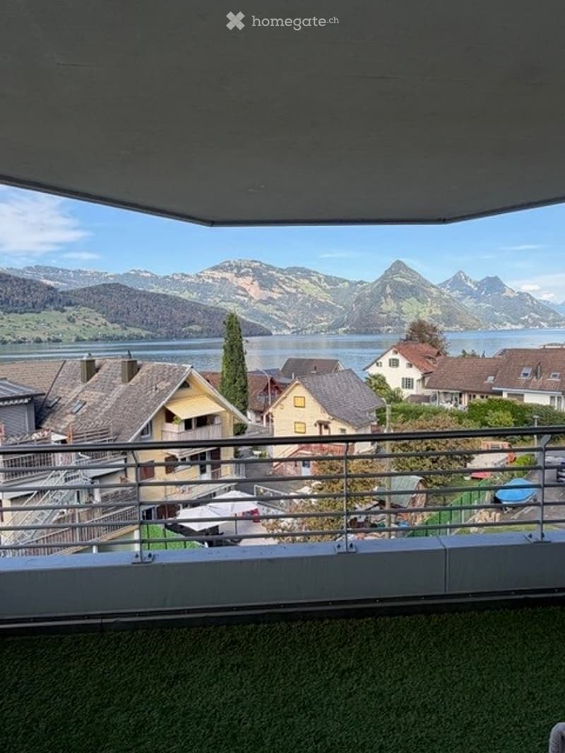 Balcony with metal railing, overlooking a scenic lake and mountain landscape, with houses and trees in the foreground.
