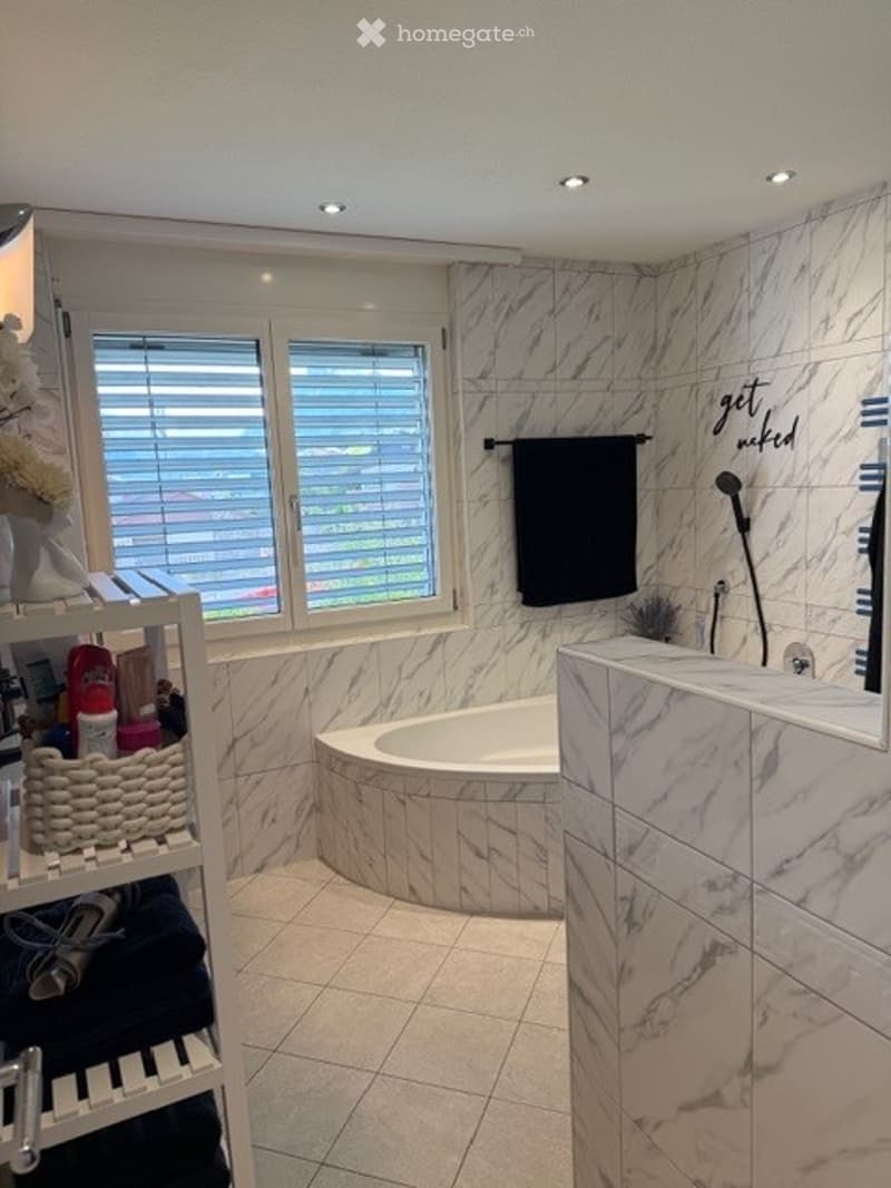 Modern bathroom with marble tiles, a freestanding bathtub, a towel rack, a shelf with various items, and a window with blinds.