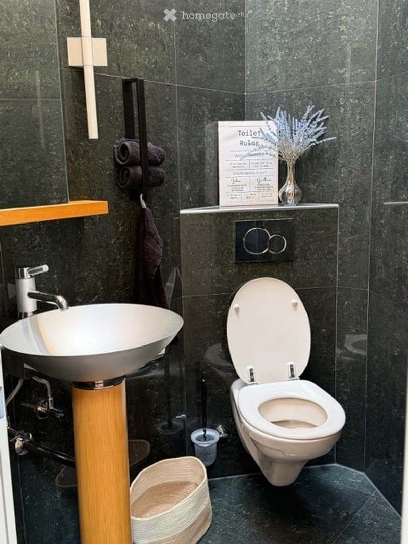 Modern bathroom with dark green tiles, a white sink with a wooden stand, a toilet, a towel rack with towels, a small shelf with a vase of flowers, and a decorative sign.