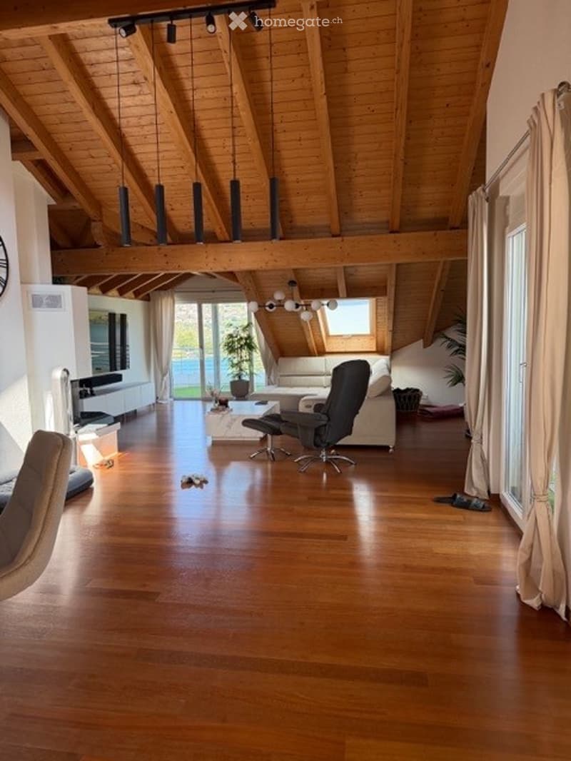 Spacious living room with exposed wooden beams, hardwood flooring, large windows, and a view of the garden. Features a white sofa, coffee table, chairs, and a television.