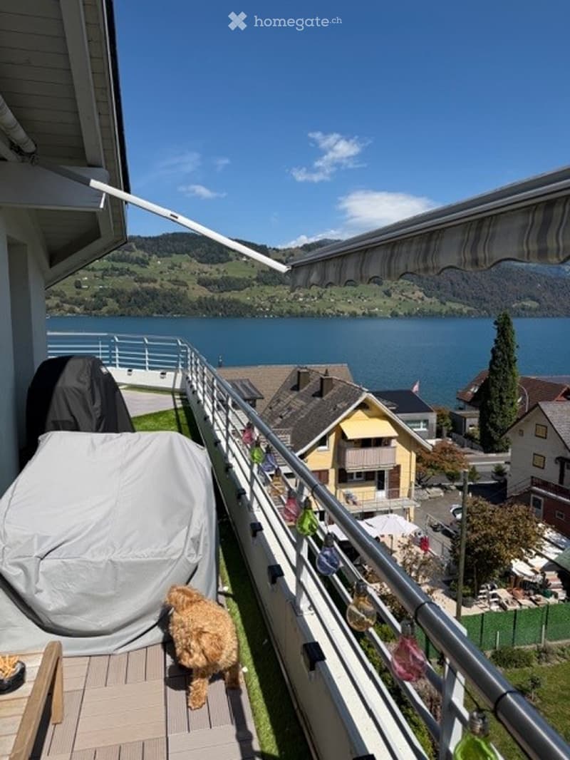 Balcony overlooking a lake with a covered grill, railing, and a small dog.