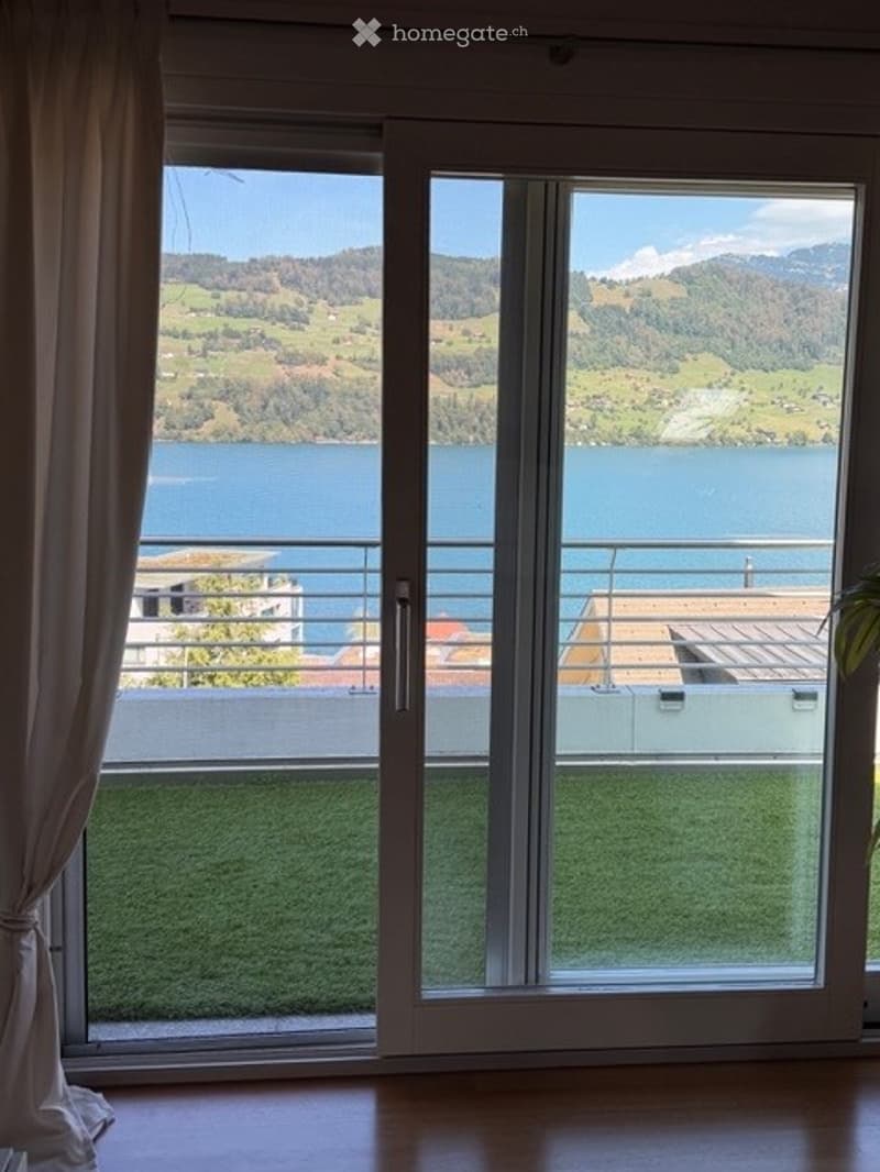 Sliding glass doors open to a balcony with a scenic lake and mountain view. The balcony has a wooden deck and a small grassy area.