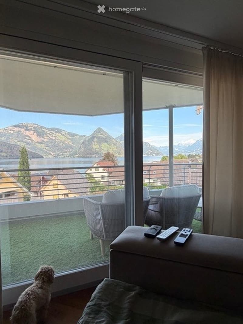 A modern living room with large windows offering a scenic mountain view, featuring a sofa, chairs, and remote controls on a small table.