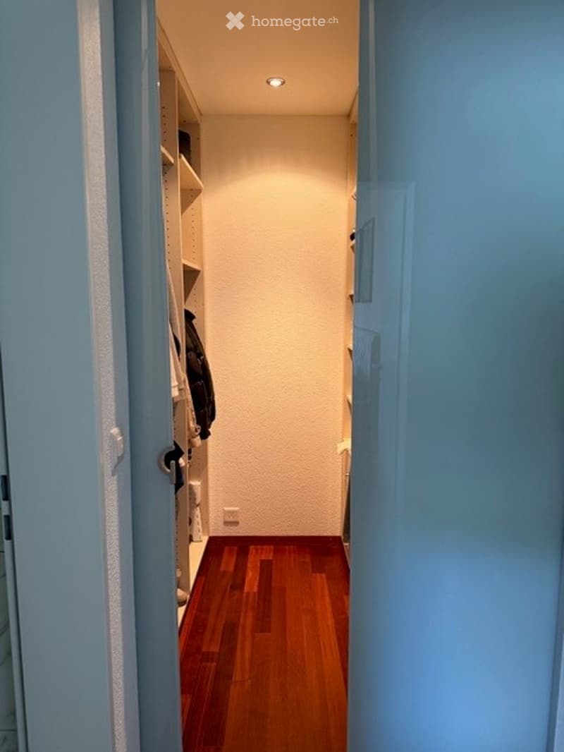 A narrow hallway with wooden flooring and white walls, featuring built-in shelving on one side and a recessed ceiling light.