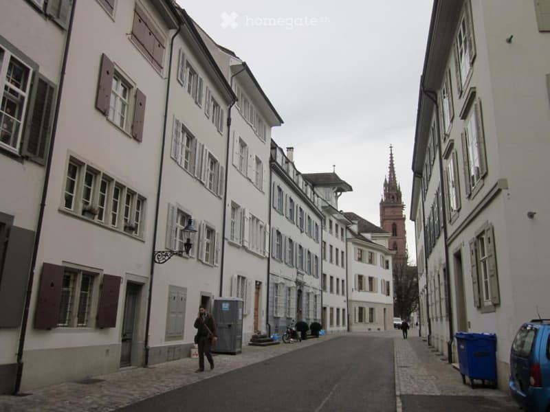 A narrow street lined with multi-story residential buildings featuring classic European architecture, with shutters and windows. A church spire is visible in the background.