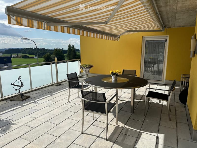Furnished balcony with table, chairs, flowers, and a decorative sundial, shaded by an awning