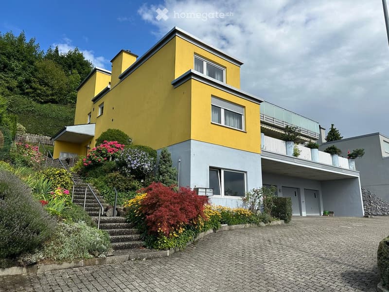 Three story house, yellow and gray exterior, balcony on second floor, brick driveway, surrounding greenery, plants on balcony