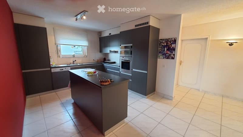 Modern kitchen, island counter, stainless steel appliances, gray cabinets, tiled flooring, window with blinds, artwork on wall, entrance door.