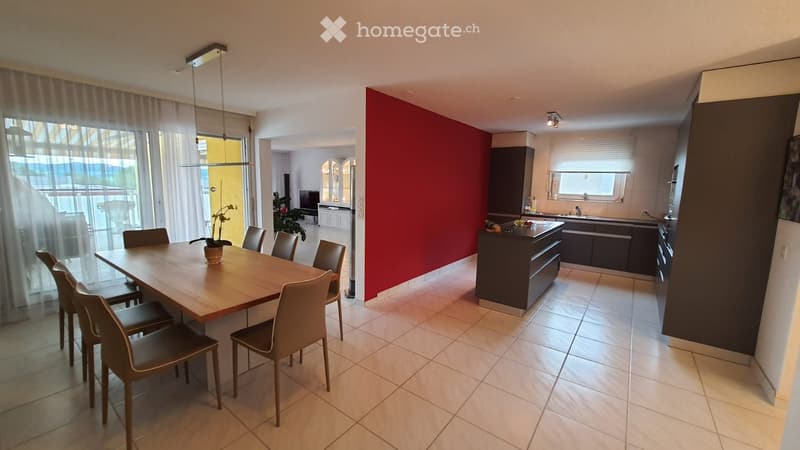 wooden dining table, wooden chairs, dining room has white tile flooring, open layout connecting with the kitchen, kitchen has island with kitchen sink and faucet, a bright red wall, wooden cabinets