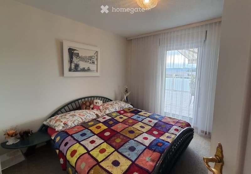 spacious bedroom with bed, decorative quilt, nightstand with figurines, framed photo, balcony with curtains, white walls, ceiling light