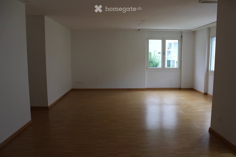 Empty room with wooden flooring, white walls, and large windows allowing natural light to enter.