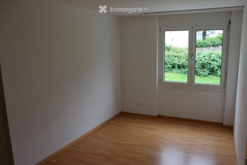 Empty room with light wooden flooring, white walls, and a large window with white frame showing greenery outside.