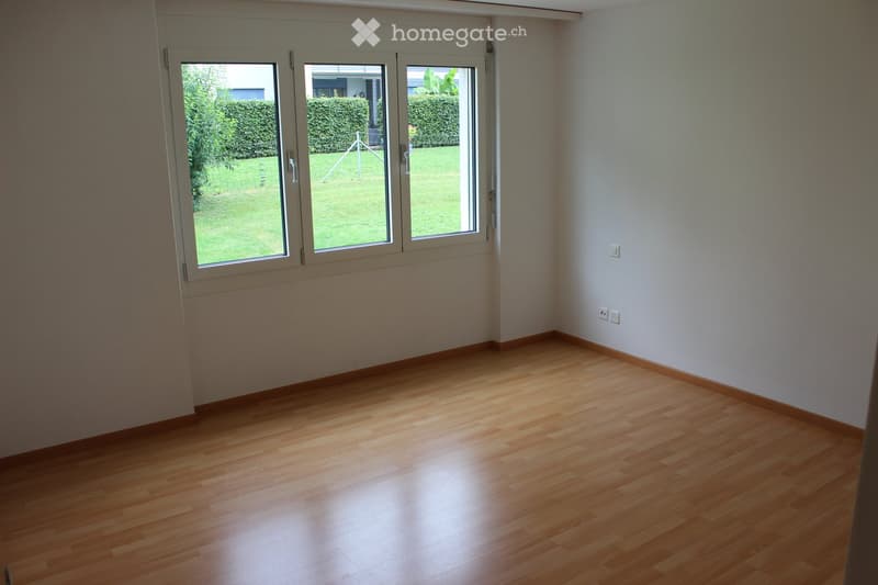 Empty room with wooden flooring and large windows leading to a garden. No furniture or equipment present.