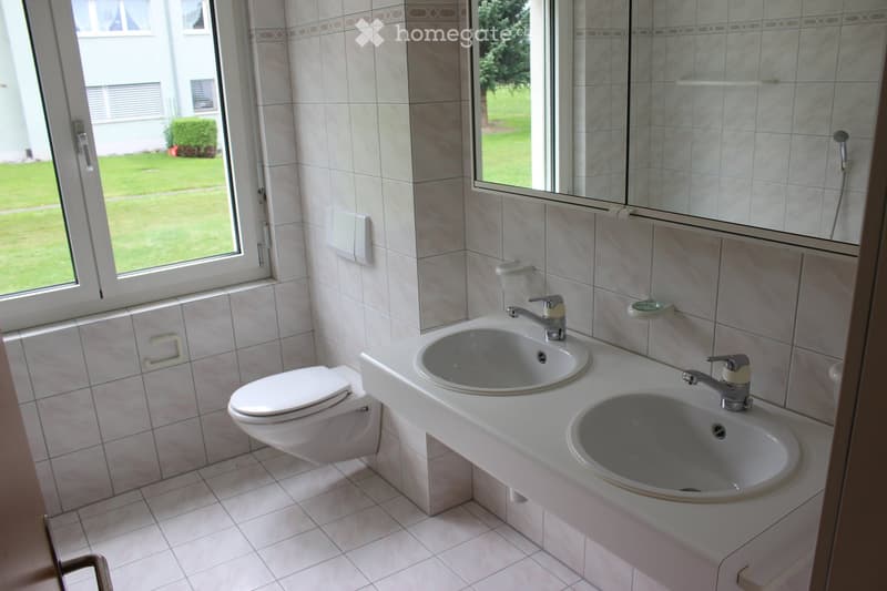 Bathroom with two sinks, a toilet, and tiled walls and floor. Large windows provide natural light and a view of the outdoor area.