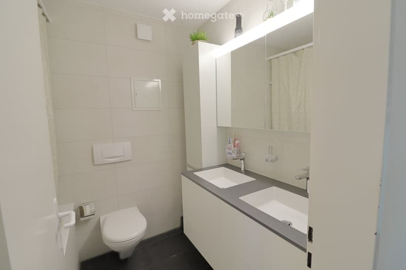 Modern bathroom with white tiles, a toilet, double sink, mirrored cabinet, and built-in lighting above the sink.