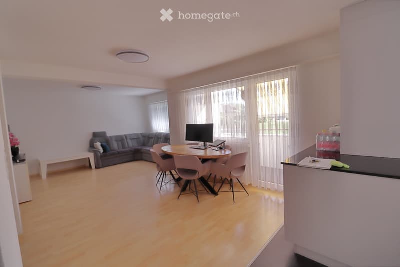 A modern living room with a grey sectional sofa, a round wooden dining table with four chairs, a flat-screen TV, and a small bench. The room features light wooden flooring and large windows with sheer curtains.