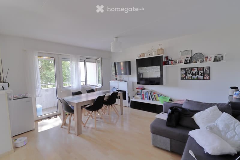 Bright living and dining area with a grey sectional sofa, a light wooden dining table with four black chairs, a television mounted on the wall, shelves with books and decorative items, and a small refrigerator near the window.