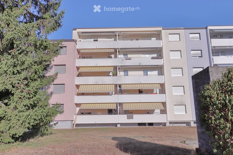 Multi-story residential building with balconies, striped awnings, and surrounding greenery.