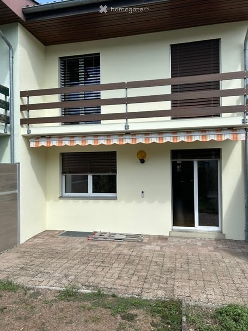 Two-story house with a balcony, beige exterior, and a covered entrance area. The ground floor has a brick patio and a small grassy area.