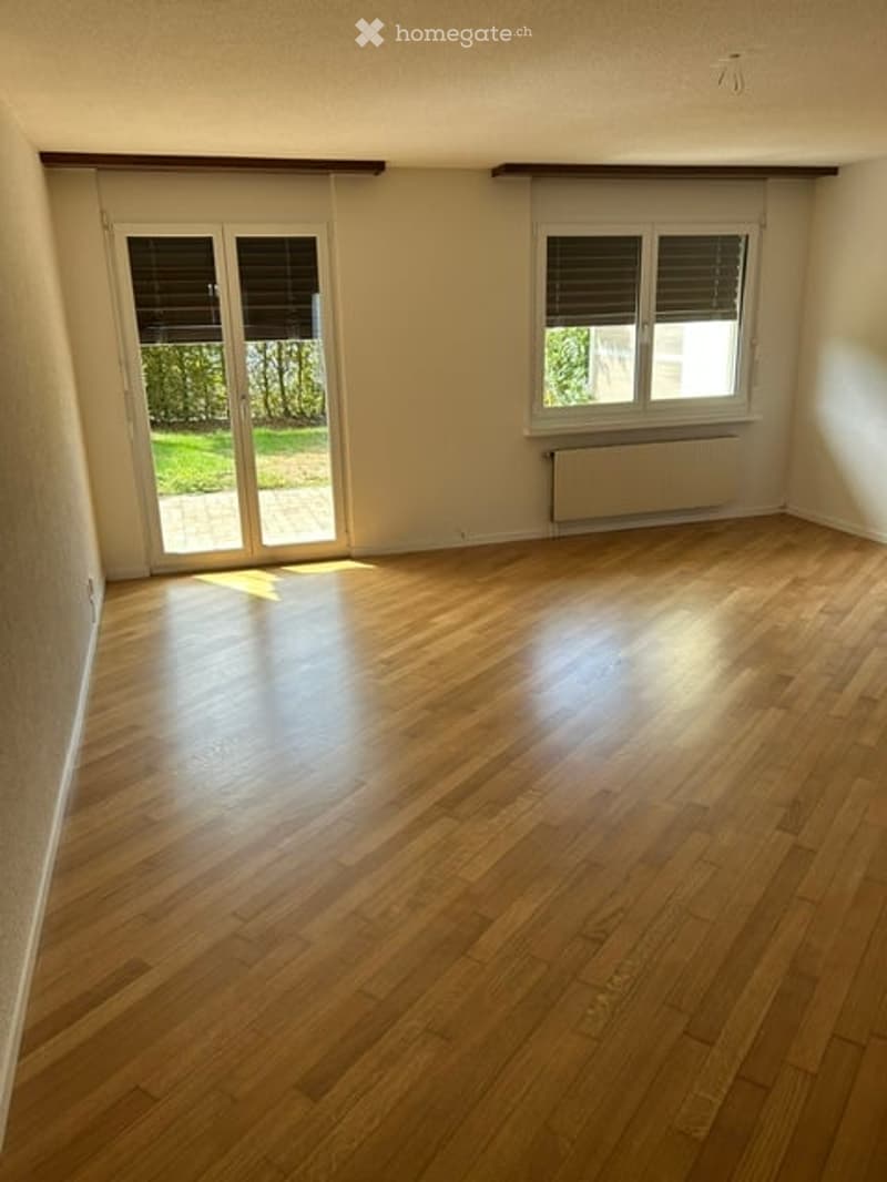 Empty room with wooden flooring, large window with blinds, and sliding glass door leading to outdoor area.