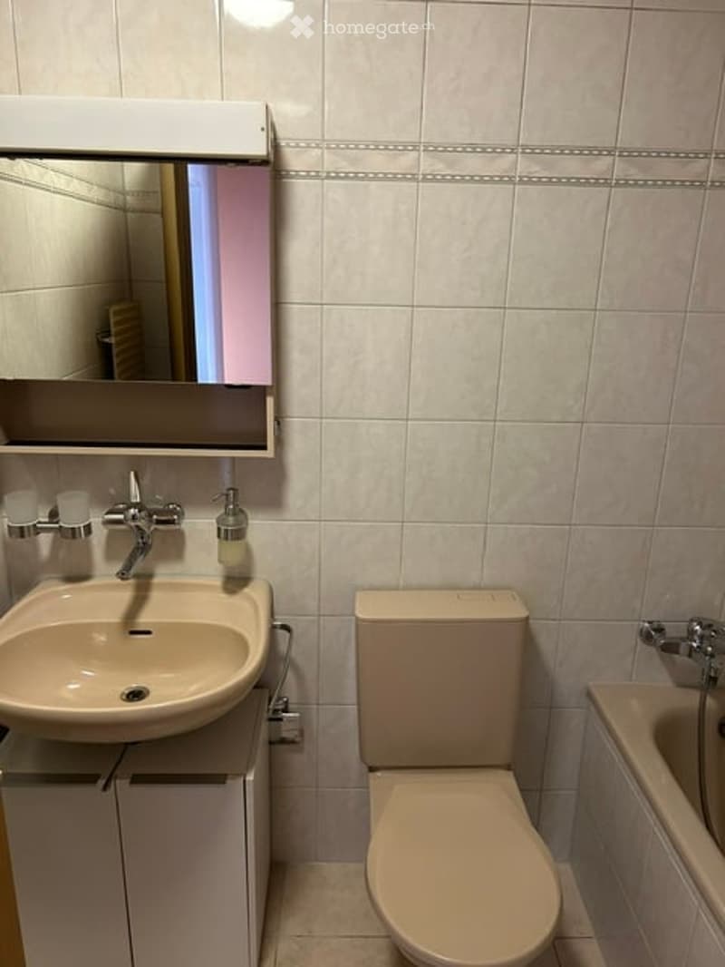 A small, clean bathroom with white tiles, a sink with a modern faucet, a mirror above it, a toilet, and a bathtub.
