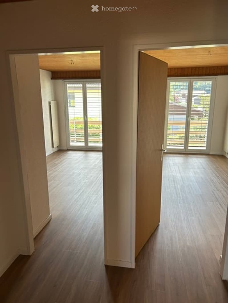 Empty room with wooden flooring, large windows, and open doors leading to another room.